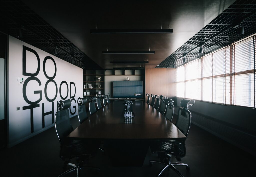 A dark conference room with a square table, black metal office chairs and a "DO GOOD THINGS" mural in the background. 