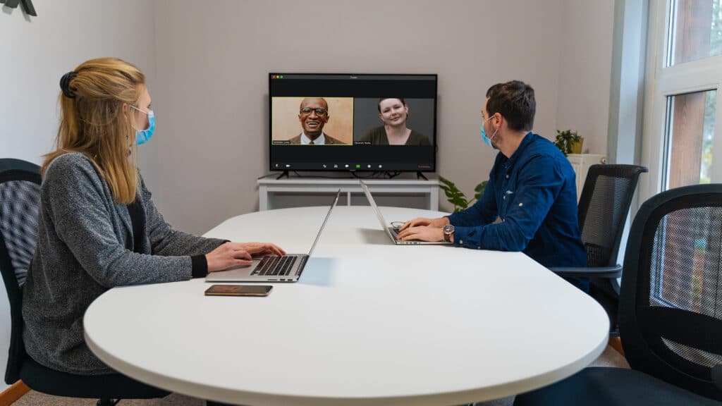 Two masked individuals are video conferencing with two colleagues via Zoom in a conference room.